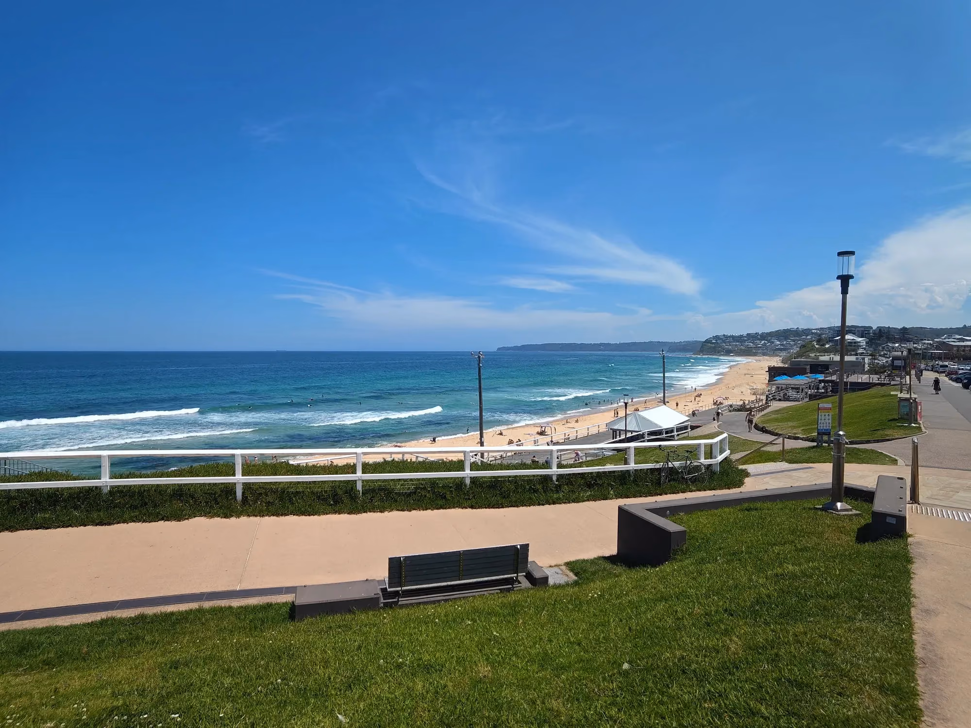A grassy clifftop path with a bench and a white rail above a long sandy beach, surfers in the waves and houses on the headland beyond